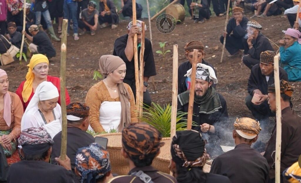 A group of people in traditional clothing sit around a central woven basket with a small plant during an outdoor ceremony as steam or smoke rises from the center